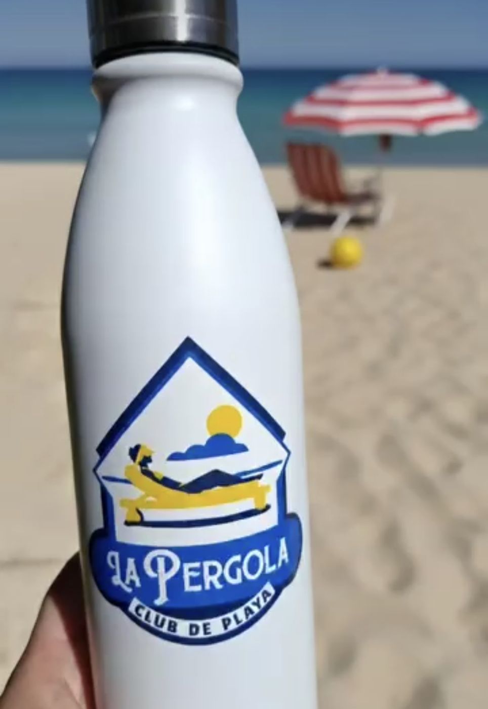 Botella blanca con logo de La Pérgola Club de Playa en la playa